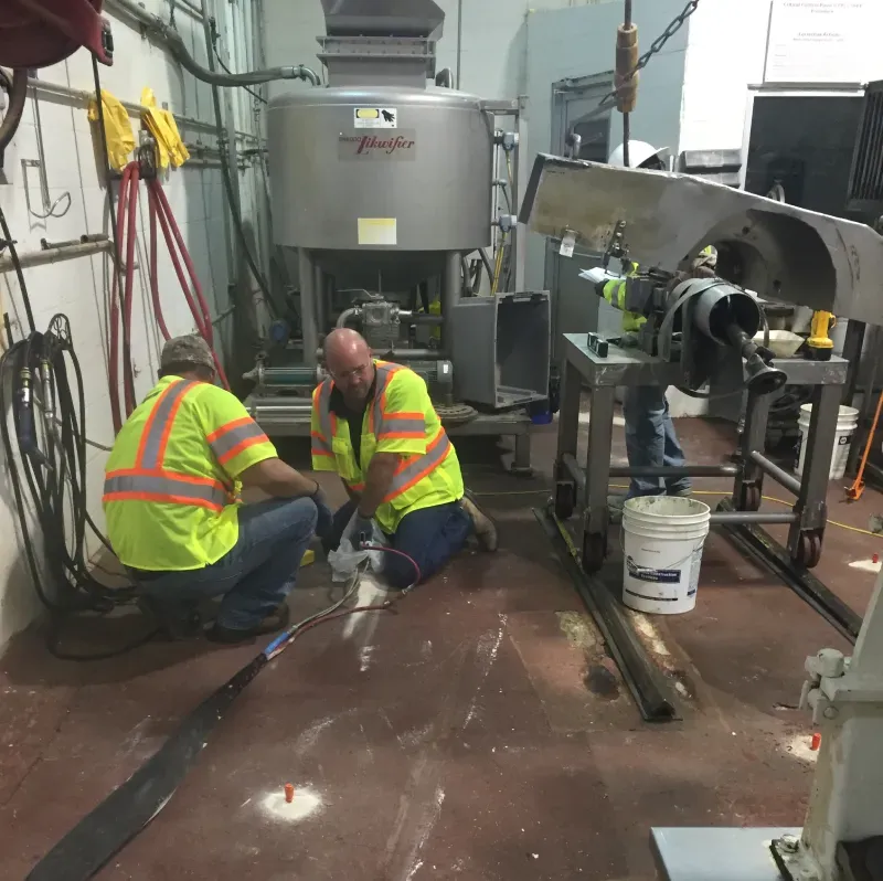 Two workers in high-vis vests doing concrete repair in machinery factory.