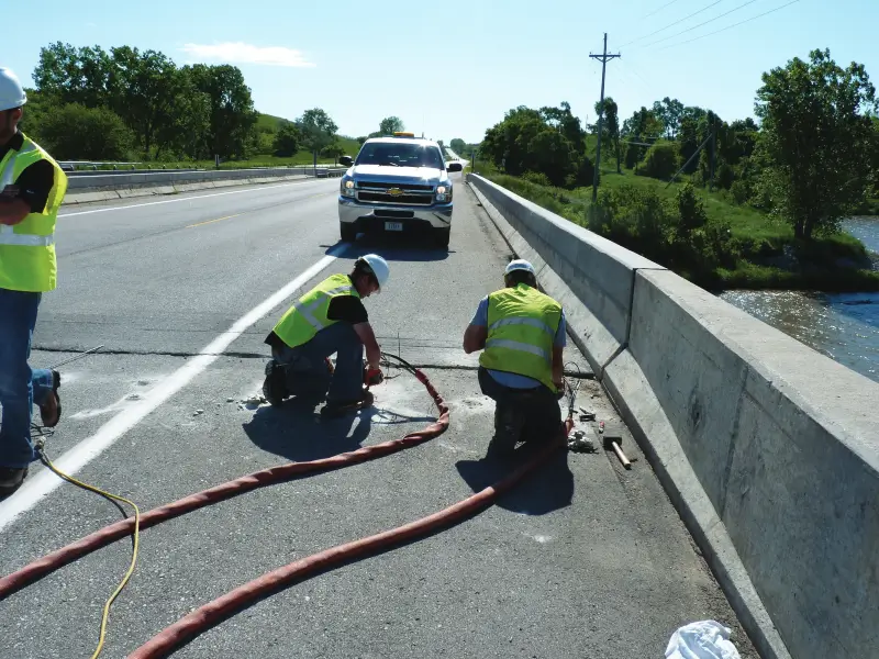 Workers repairing road on sunny bridge with vehicle nearby.